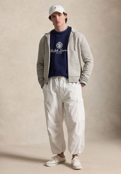 Jeune homme portant une casquette blanche, un sweat Ralph Lauren bleu marine, un sweat à capuche gris clair zippé, un pantalon cargo blanc et des baskets blanches sur fond uni.