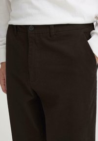 Pantalones chinos de algodón marrón con una textura suave, con cierre de botón y bolsillos laterales, parcialmente visibles sobre un fondo claro.