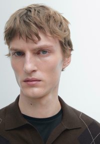 Maglia polo marrone lavorata a maglia con colletto, caratterizzata da un motivo geometrico. Il modello ha capelli corti e spettinati e uno sfondo neutro.