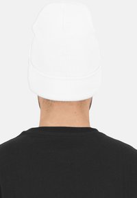 Persona che indossa un semplice cappellino lavorato a maglia bianco e una maglietta nera, vista da dietro su uno sfondo grigio chiaro.