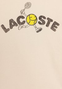 Crème katoenen t-shirt met een cartooneske gele tennisbal met een lach, die een racket vasthoudt, en de tekst "LACOSTE" in vetgedrukte zwarte letters.
