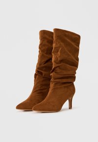 Bottes en daim marron arrivant au genou avec un bout pointu et un talon effilé, présentant un design drapé et une finition texturée.