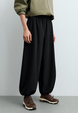 Personne portant un pantalon noir ample avec des poignets élastiques, associé à des chaussures de randonnée marron et une manche de veste vert olive visible.