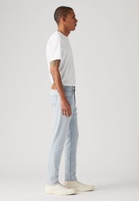 Ανοιχτό μπλε τζιν με στενή εφαρμογή, φορεμένο με απλό λευκό T-shirt με στρογγυλή λαιμόκοψη και λευκά αθλητικά παπούτσια, σε πλευρική όψη.