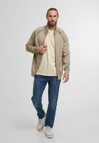 Beige zip-up jacket with ribbed cuffs and collar, paired with a light t-shirt and blue jeans, worn with white and cream sneakers.