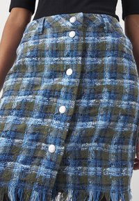 Jupe à carreaux bleue et verte avec un tissage texturé, ornée de boutons argentés sur le devant et d'un bord inférieur à franges.