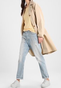 Donna che indossa un trench coat beige, una maglietta a righe gialle, jeans chiari strappati e sneaker bianche, in piedi con una gamba in avanti su uno sfondo bianco.