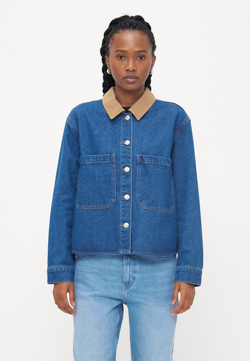 Casaco de ganga com um colarinho em corino castanho claro, bolsos frontais e botões prateados. Estilizado com uns jeans de cor azul clara, apresentando uma textura suave.