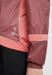 Vindtæt jakke med et bordeaux yderlag og lyserøde mesh-sidelameller, med elastiske manchetter og refleksdetaljer for synlighed.