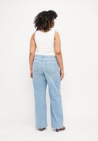 Jeans azul claro de pernas largas com uma cintura média, apresentam bolsos traseiros e um detalhe de costura discreto. Combinados com uma t-shirt branca.