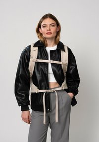 Veste bomber en cuir noir avec un harnais beige, des poignets côtelés et une taille à cordon. Associée à un pantalon gris et un haut blanc.