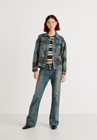 Jeans jakna s cvetličnim potiskom, pod njo krajša striped pulover, v kombinaciji z razširjenimi modrimi džinsami in črnimi čevlji s špičastim nosom.