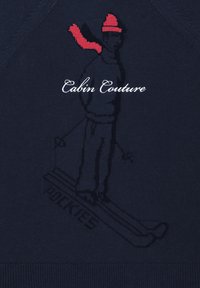 Laivastonsininen villapaita, jossa neulottu hiihtäjäkuvio, punainen hattu ja huivi. Ylhäällä teksti "Cabin Couture" ja alhaalla hienovaraisella tekstuurilla "POCKIES".