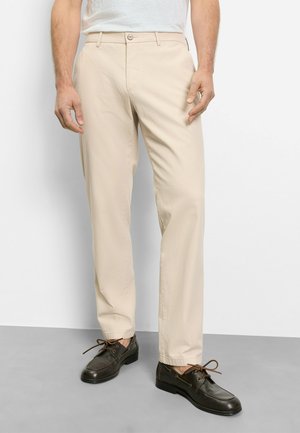 Man die beige broek en donkerbruine leren veter schoenen draagt, staand op een grijze vloer tegen een witte muur.