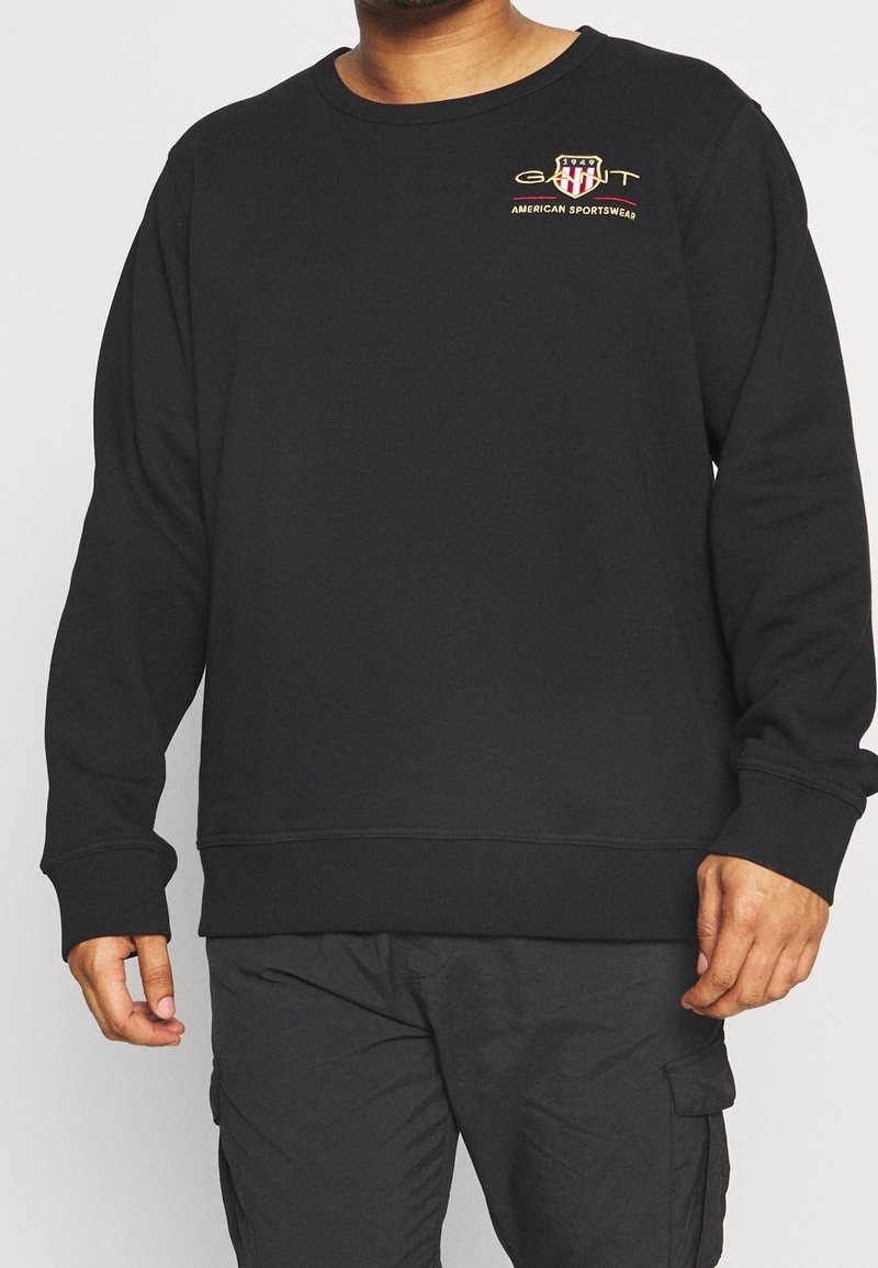 Homme portant un sweat-shirt noir avec un petit logo "GANT 1949 American Sportswear" sur le côté gauche de la poitrine, associé à un pantalon cargo noir.