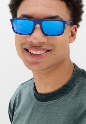 Giovane persona sorridente con occhiali da sole rettangolari specchiati blu e maglietta verde a coste con collo rotondo.