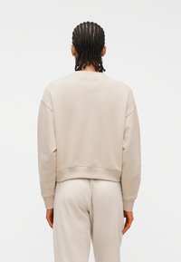 Άτομο με πλεκτά μαλλιά που φοράει μπεζ μακρυμάνικη μπλούζα και αντίστοιχο παντελόνι, φωτογραφημένο από πίσω σε απλό λευκό φόντο.