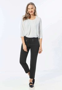 Pantalon noir avec poches zippées ; associé à un chemisier blanc plissé. Le modèle a de longs cheveux gris et porte des chaussures navy. Design simple et coupe confortable.