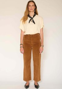 T-shirt crème avec une coupe décontractée, associé à un pantalon en velours côtelé marron et des ballerines noires ornées de clous en argent, accentué par une écharpe noire.