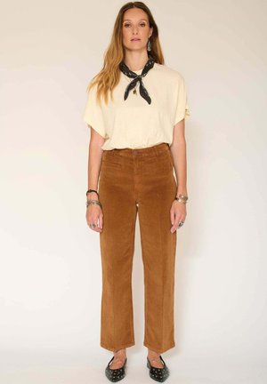 T-shirt crème avec une coupe décontractée, associé à un pantalon en velours côtelé marron et des ballerines noires ornées de clous en argent, accentué par une écharpe noire.