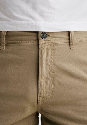 Beige bukser med knap og lynlås, frontlomme, bæltestropper, båret med en hvid skjorte, der er synlig ved taljen.