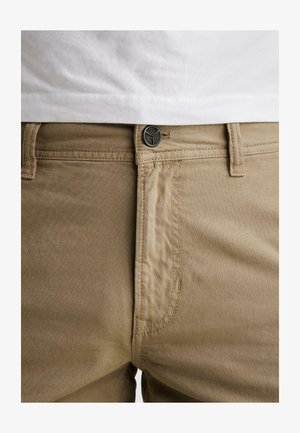 Beige bukser med knap og lynlås, frontlomme, bæltestropper, båret med en hvid skjorte, der er synlig ved taljen.