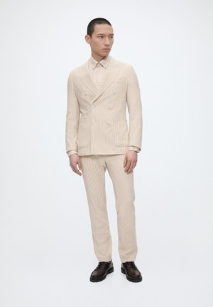 Mężczyzna ubrany w beżowy garnitur dwurzędowy z dopasowanymi spodniami i ciemnobrązowymi butami stoi na jednolitym białym tle.