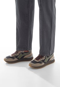 Gambe che indossano pantaloni grigio scuro e sneakers nere, beige e verde oliva con suola in gomma su uno sfondo bianco.