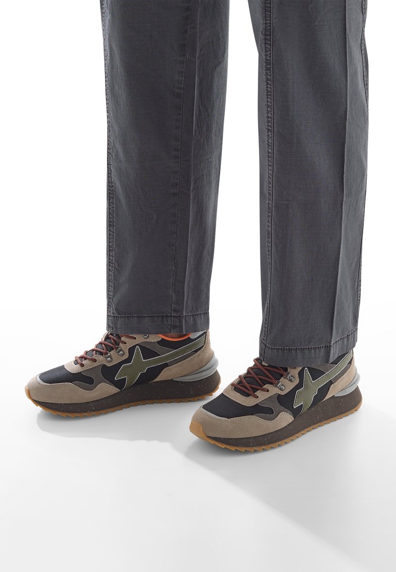 Gambe che indossano pantaloni grigio scuro e sneakers nere, beige e verde oliva con suola in gomma su uno sfondo bianco.
