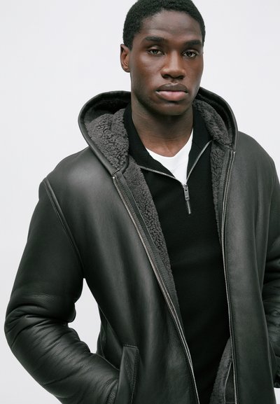 Schwarze Lederjacke mit Kapuze und grauem Fleece-Futter, ausgestattet mit einem Reißverschluss und zwei Vordertaschen, präsentiert auf einem neutralen Hintergrund.