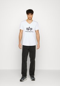 Bílý tričko s výstřihem do V a černým logem "Alpha Industries", v kombinaci s černými kalhotami a šedými teniskami, stojící na neutrálním pozadí.