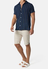 Niebieska koszula w paski z krótkim rękawem, z białymi guzikami, w połączeniu z beżowymi szortami z wywijanym dołem i białymi sandałami z podwójną klamrą.