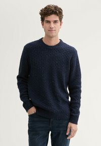 Donkerblauwe gestructureerde gebreide trui met een ronde hals, voorzien van een gespikkeld patroon en geribbelde manchetten en zoom. Gekleed met donkere jeans.