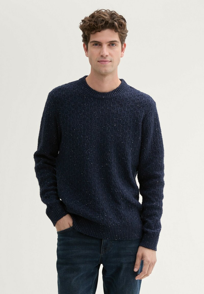 Donkerblauwe gestructureerde gebreide trui met een ronde hals, voorzien van een gespikkeld patroon en geribbelde manchetten en zoom. Gekleed met donkere jeans.