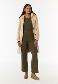 Beige wollen jas met een capuchon, olijfgroene ribgebreide top en bijpassende wijde broek, gecombineerd met tan veterlaarzen. Geen patronen of hardware details.