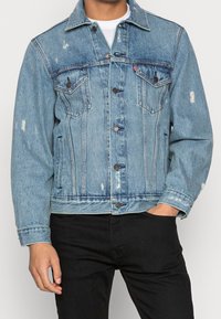 Man i ljusblå denimjacka med knäppning fram, slitna detaljer och svarta byxor, med handen i fickan mot en enfärgad bakgrund.