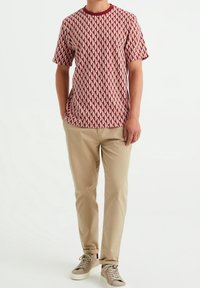 Kortärmad skjorta med ett rött geometriskt mönster, rund halsringning och en avslappnad passform, kombinerad med beige byxor och lätta sneakers.