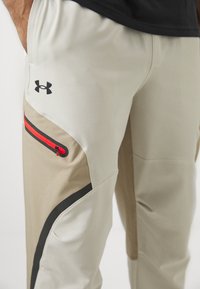 Pantalons de sport pour hommes en beige clair avec des accents noirs. Équipés d'une poche zippée avec une fermeture éclair rouge et un logo noir sur la cuisse.