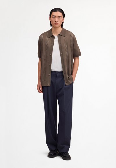 Jeune homme portant un cardigan marron à manches courtes boutonné par-dessus un t-shirt blanc, un pantalon large bleu marine et des chaussures noires, debout avec une main dans la poche.