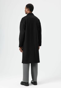 Schwarzer Wollmantel mit klassischem Kragen, langen Ärmeln und geradem Schnitt, kombiniert mit grauer Hose und schwarzen Schuhen. Schlichter Hintergrund.