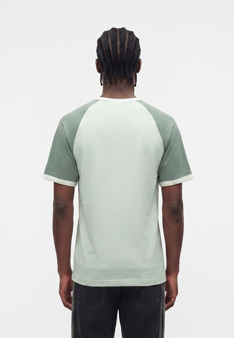 Mand med cornrow fletninger iført en lysegrøn og hvid raglan T-shirt, stående med ryggen mod en ensfarvet baggrund.