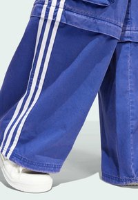 Blå, baggy bomullbyxor med vita sidosträck. Har vida ben och en fåll som vilar ovanför vita sneakers.