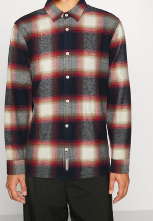 Mężczyzna noszący koszulę z długim rękawem w czerwono-czarno-biało-szary wzór w kratę, w połączeniu z czarnymi spodniami.