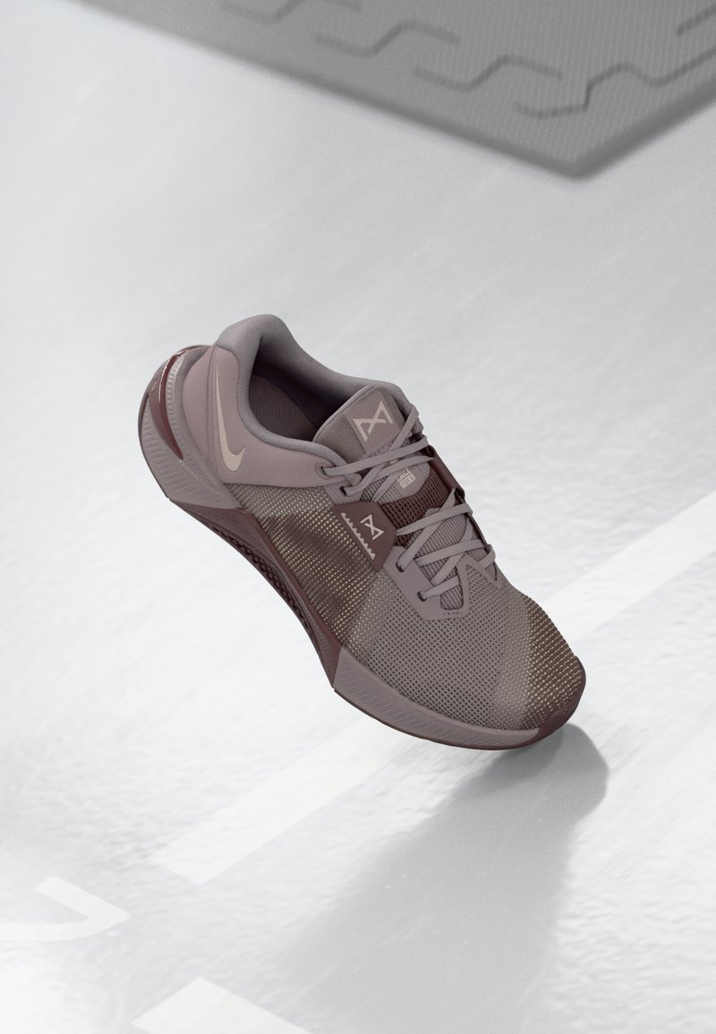 Zapatilla deportiva gris claro con materiales de malla y sintéticos, que presenta una suela texturizada y punta redonda, acentuada por detalles y un logo en gris más oscuro.