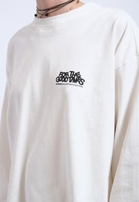 Langärmliges, cremefarbenes Baumwoll-T-Shirt mit einem schwarzen Grafikdruck, der "FÜR DIE GUTEN ZEITEN" sagt. Einfaches und entspanntes Design.