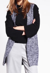 Vrouw met gekruiste armen in een zwart geribbeld topje, een grijze gestructureerde cardigan met een open voorkant en lichtgrijze broek tegen een witte achtergrond.