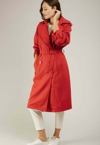 Manteau trench rouge avec taille ceinturée, grand col, boutons double boutonnage et poches latérales, associé à un pantalon blanc et des baskets.