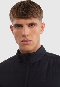 Juodas zip-up džemperis su aukštu apykaklės kraštu, pagamintas iš lygaus audinio. Su subtiliu tekstūruotu raštu ir logotipu ant krūtinės.