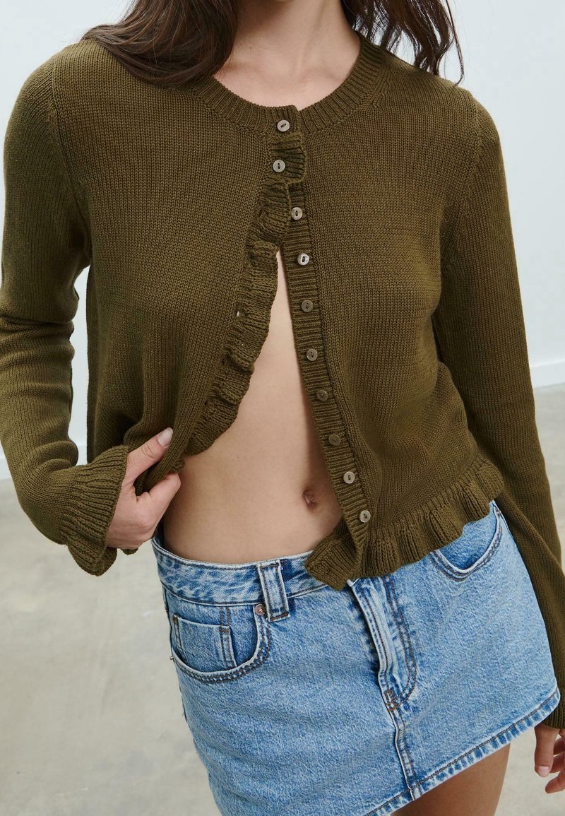 Femme portant un cardigan vert olive boutonné partiellement ouvert, assorti à une mini-jupe en denim bleu clair taille haute.