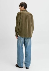 Persona in piedi di spalle, con indosso una camicia verde oliva oversize, jeans chiari larghi e scarpe scure, su uno sfondo bianco semplice.